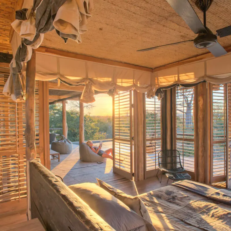 Beautiful khaki-themed safari lodge room with cozy couches, overlooking the African wilderness, with a guest reading outside at Asilia Lodge in Tanzania.