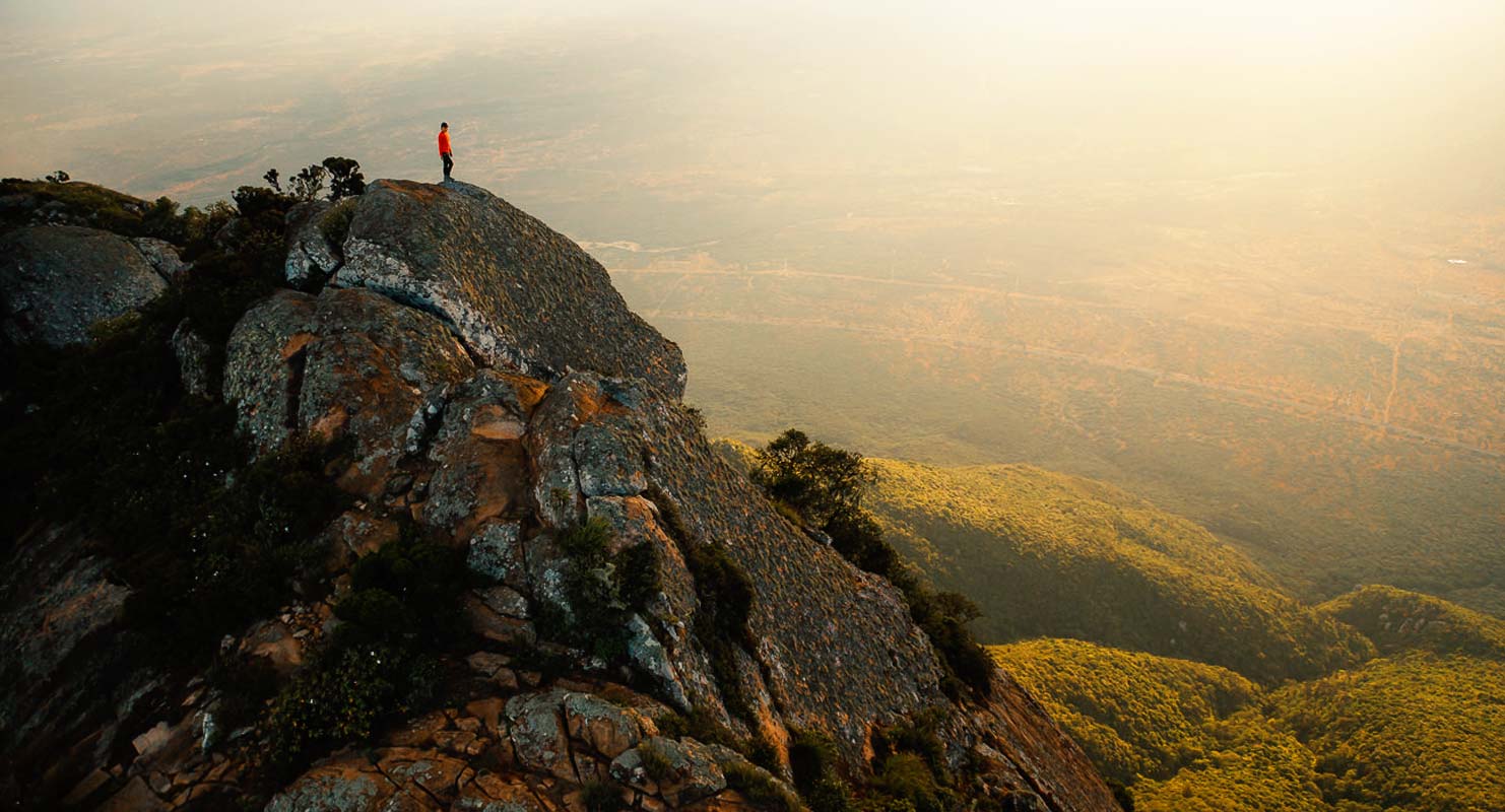 A hiker stands triumphantly atop a peak of Longido Mountain, gazing over the expansive Tanzanian landscape with Mount Kilimanjaro visible in the distance.