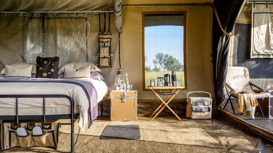 Cozy safari lodge room with a comfortable bed, wooden chair, and a table set with refreshing drinks, creating the perfect relaxation space in a luxury safari camp.