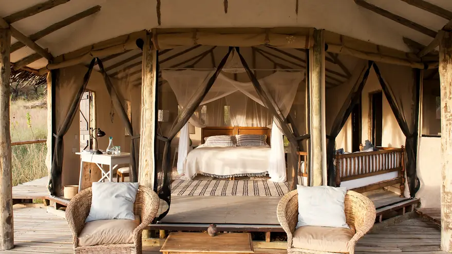 Stunning luxury safari lodge bedroom with a comfortable bed and two lovely chairs, elegant furnishings, and a serene ambiance for a perfect Tanzania safari stay.